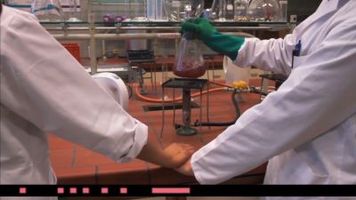 Film still from 'Touching the Cheese' (2017) by Jon Beaton. Two scientists in lab coats stand around a Bunsen burner holding hands while one of them swirls a conical flask over the burner with their free hand.